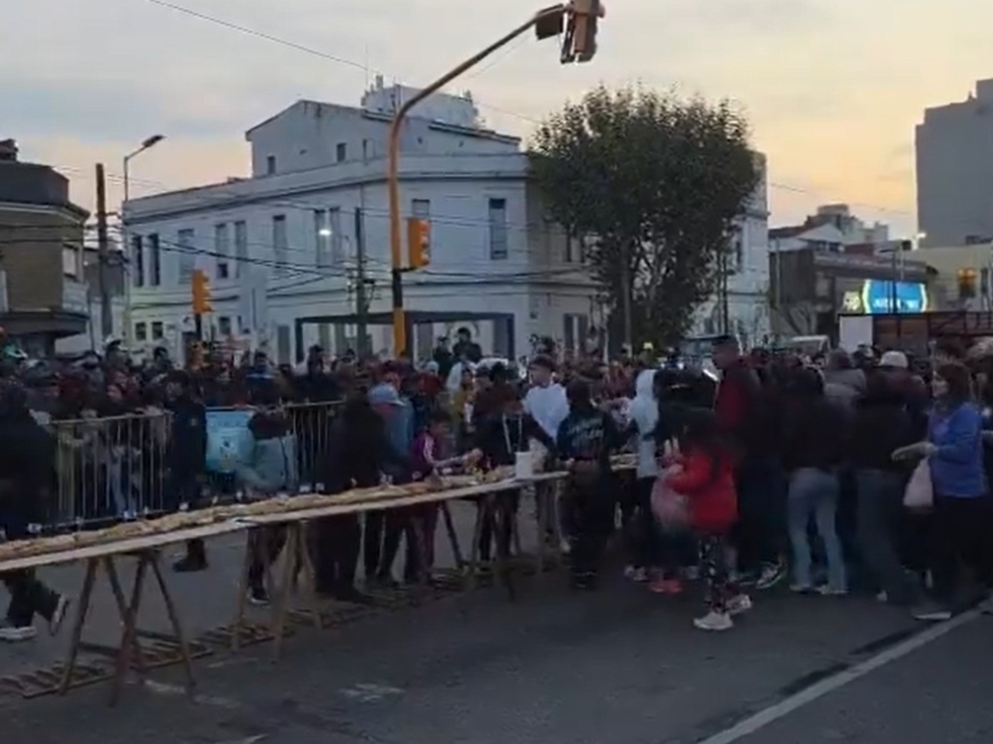 A crowd of people pushing and grabbing at a long sandwich laid out on tables after breaching barriers.