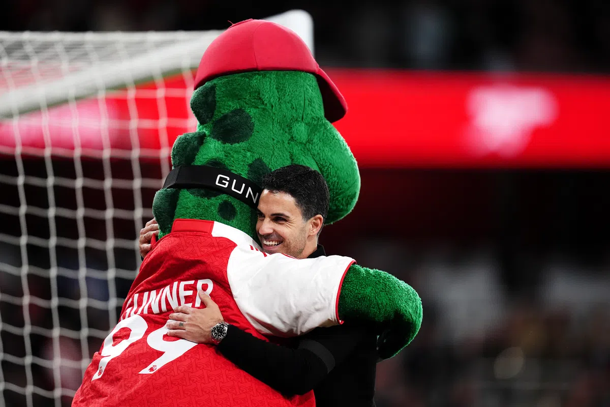 Mikel Arteta raising his arms in celebration on the pitch, surrounded by Arsenal players and fans, with confetti falling.