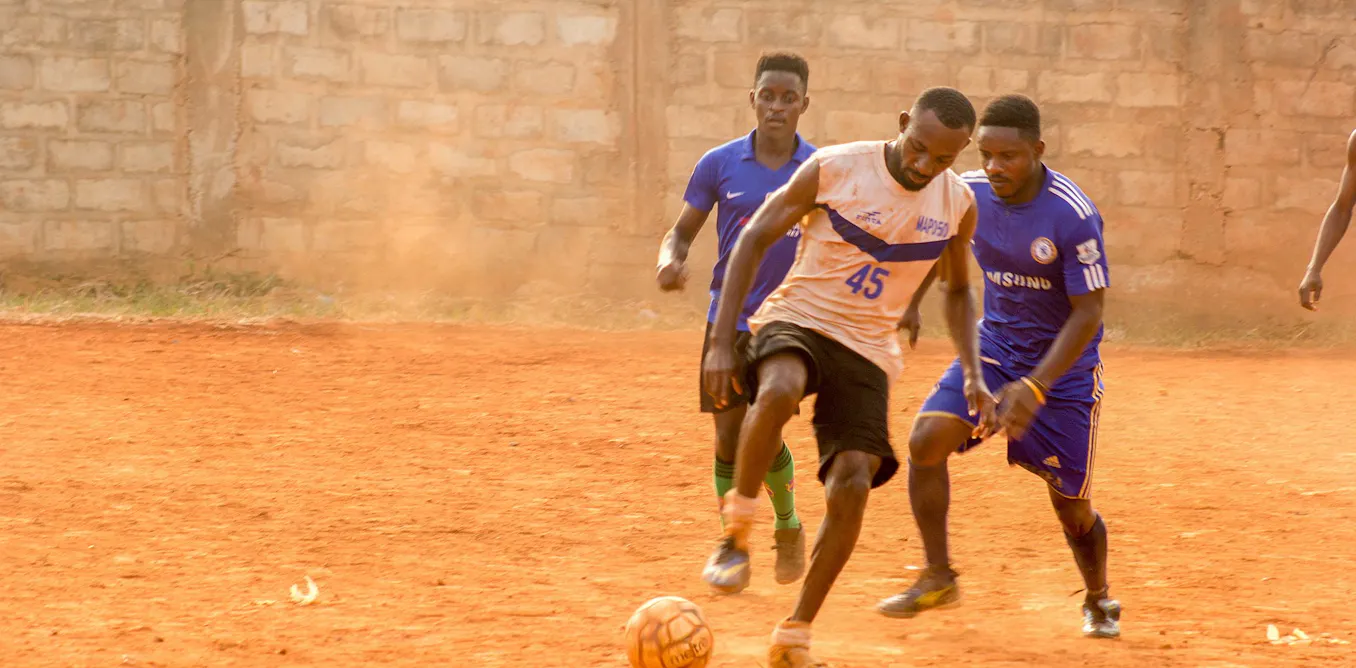 Young Ghanaian men playing football on a dusty pitch, symbolizing their dreams, with a subtle overlay hinting at digital deception.