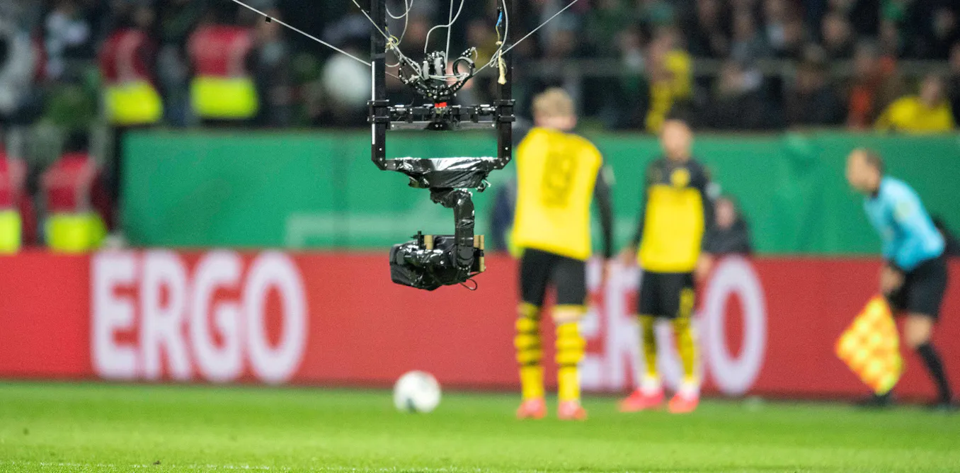 A modern football stadium with multiple advanced broadcast cameras, including a spider camera overhead, capturing a match during a major tournament.